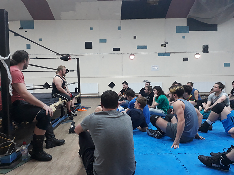 A group of men are sat on the floor around a man who is sat on a wrestling ring