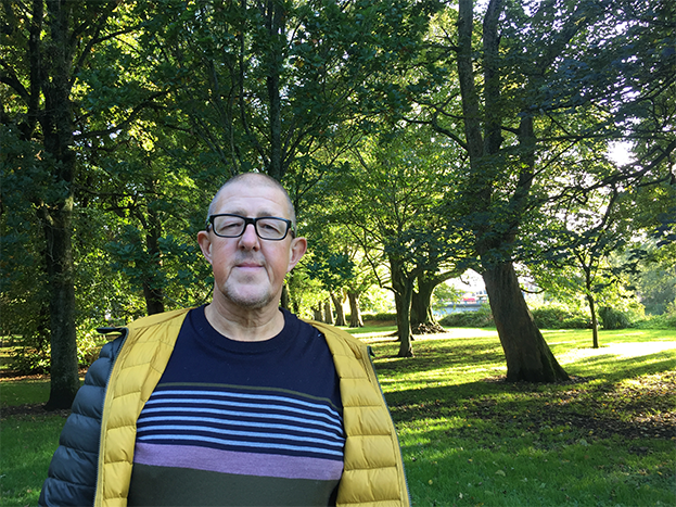 Man in yellow jacket with glasses in park on a sunny day in front of green autumnal trees