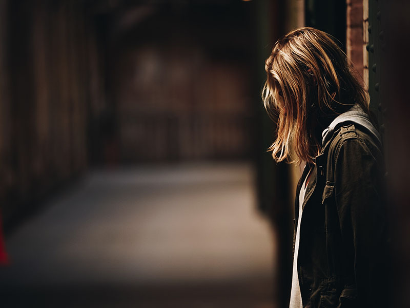 Woman looks down, her hair covers her face, the background is blurred and dark