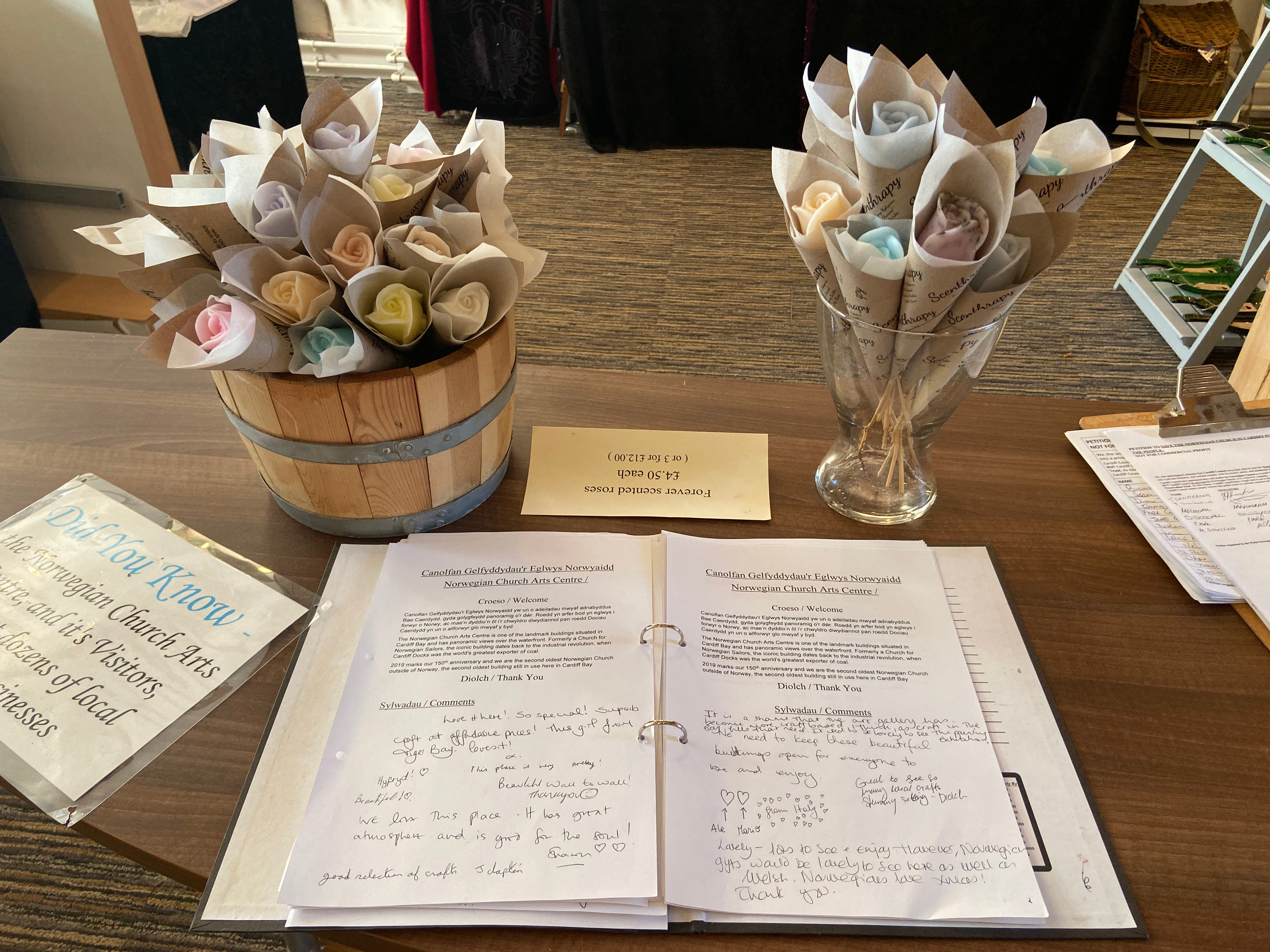 Vases of wax roses sit behind a visitor book on a wooden table. The book contains scrawled comments from visitors to the Norwegian Church Arts Centre