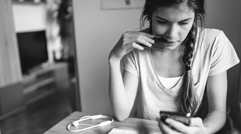 Young woman with braided hair sitting by the table, looking on her smart phone. Paying bills on the phone.